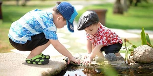 Two young boys squatting down, exploring a pond in a park.