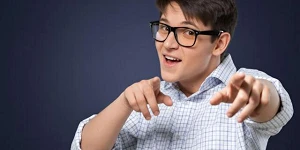 A young man with dark hair and glasses in a dress shirt, both hands pointing toward us and a smile like he just "got" us.