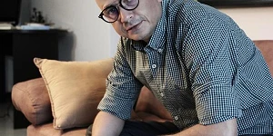 A man sitting on a couch, sleaves rolled up, book in hand, glasses on, looking directly toward us.