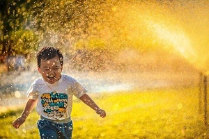 A young boy running through a sprinkler in his clothes, joy bursting from his expression.