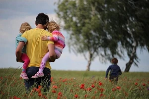 A father carrying 2 of his children through a field of long grass & poppies while a third child runs ahead.