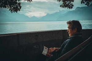 I Had a Thought But I Can’t Remember It An AI generated image of a man sitting on a bench with closed book, looking out at a calm body of water with mountains in the distance.
