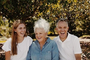 An image of 3 people from 3 generations sitting down on a bench at a park and laughing.