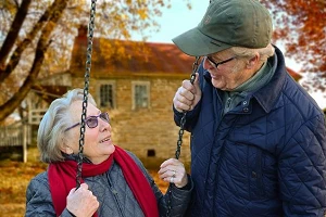 An elderly couple outdoors, she's sitting on a swing and he is standing beside her, they're looking at each other happily.