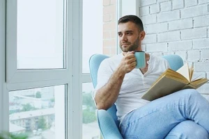 A middle-aged man wearing white t-shirt & blue jeans, holding a cup of coffee and an open book on his lap.