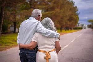An elderly couple walking arm in arm away from us along a tree-lined roadway.