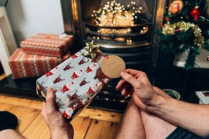 A Christmas gift in the hands of a man looking at the tag with a fireplace and more gifts in the background.