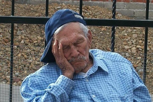 An old man, baseball cap on sideways, head resting in his hand, outdoors and fast asleep.