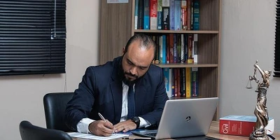 A balding man in a suit, laptop open in front of him, writing on a paper at the long table he is sitting at.