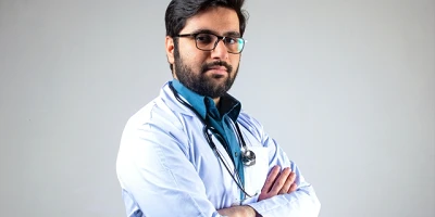 A brown skinned doctor standing in a lab coat with a stethoscope around his neck,, arms crossed, looking directly at us.