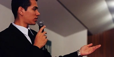 A young pastor with dark hair and wearing a dark suit and tie, mic in hand, other hand outstretched - mid-sermon.