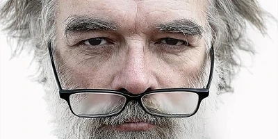 An older man with frontal baldness, glasses under his nose, looking at us grumpily.