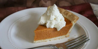A piece of pumpkin pie on a white plate with a dollop of whipped cream on top and a fork on the side.