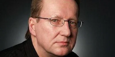 A middle-aged man with brown hair, small glasses, looking somber.
