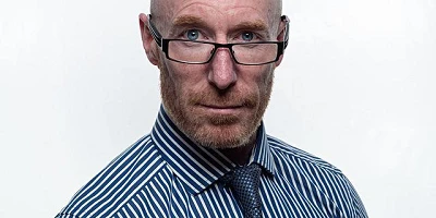 A bald man with glasses in a blue striped shirt & tie looking directly at us.