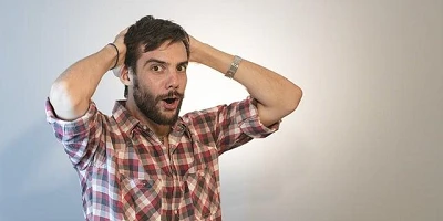 A young man in a short-sleeved red & brown checked shirt, hands on the back of his head looking alarmed.