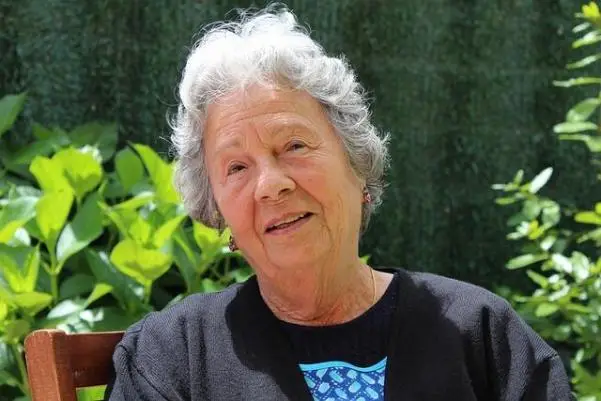 An elderly woman in casual clothes sitting on a chair outdoors.