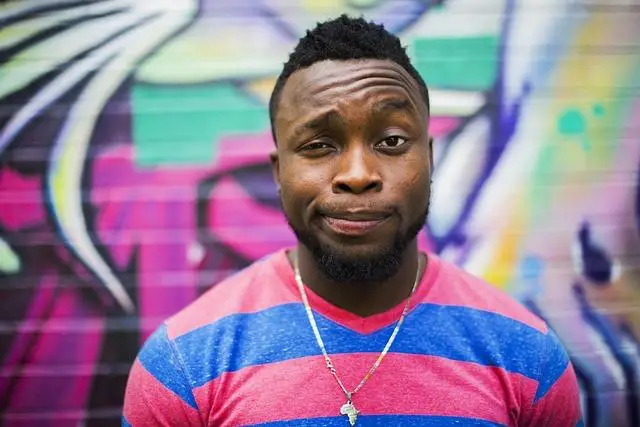 A black man in a pink and blue wide-striped shirt, one eyebrow raised.