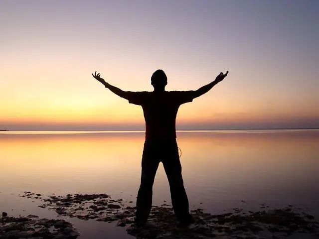 silhouette of a man standing by a lake with his hands stretched up to the sky.