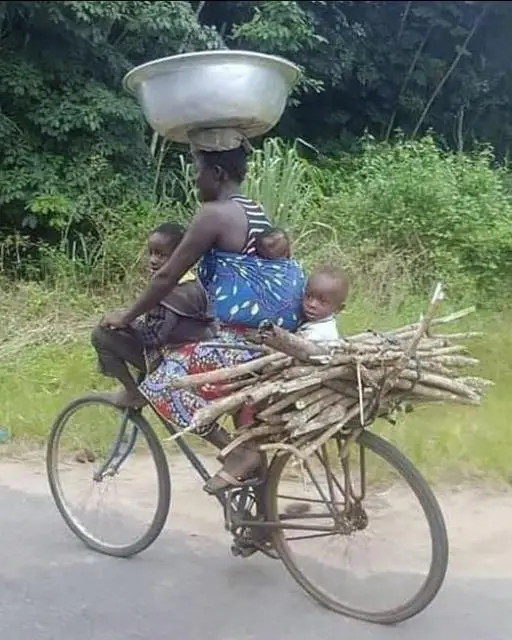 An African woman on a bicycle with a large metal basin on her head, 3 children, and a bundle of sticks.
