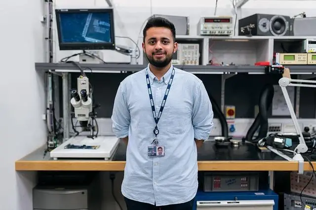 A young man in a blue shirt, wearing a lanyard with ID, standing in front of his work desk complete with all his engineering tools.