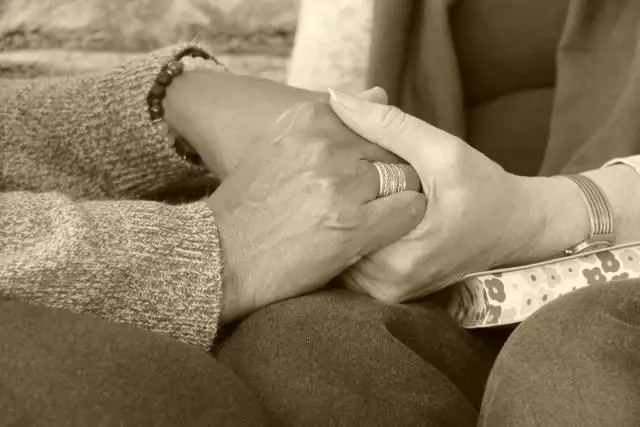 A close-up, sepia-toned photo of 2 women clasping hands, knees touching.