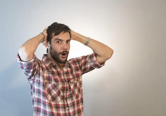 A thirty-something man with beard & hands behind his head looking very surprised.