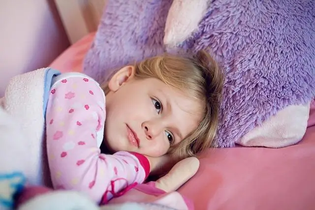 A young girl with her head on a pillow, covers up and eyes open.