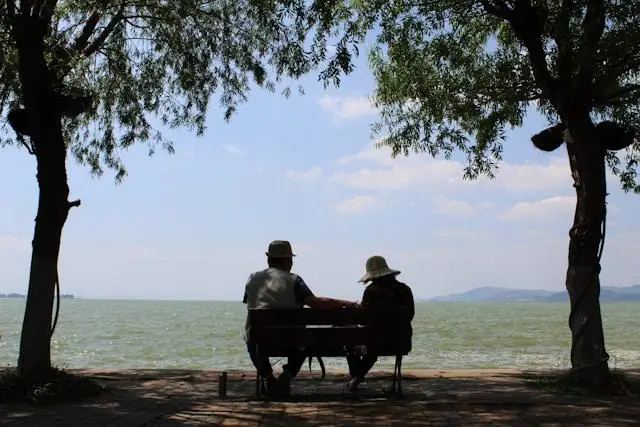 An elderly couple sitting on a park bench looking out over a lake.