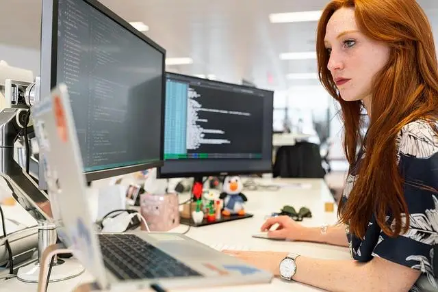 A side view of a woman in an office at a desk looking at one of her 3 computer screens.