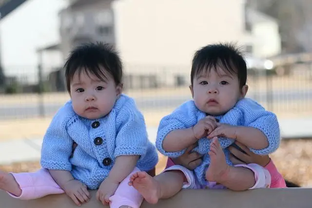 Identical twin babies dressed the same sitting on an outdoor table looking directly at us.