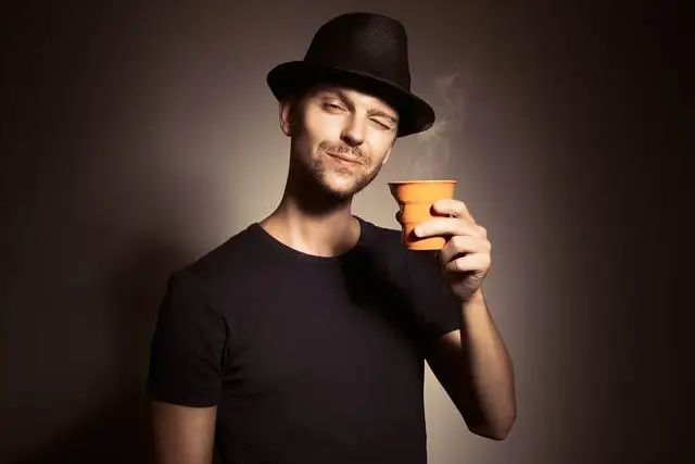 A man wearing a hat, standing in front of a blank wall, coffee cup raised, head cocked to one side with a self-satisfied expression.