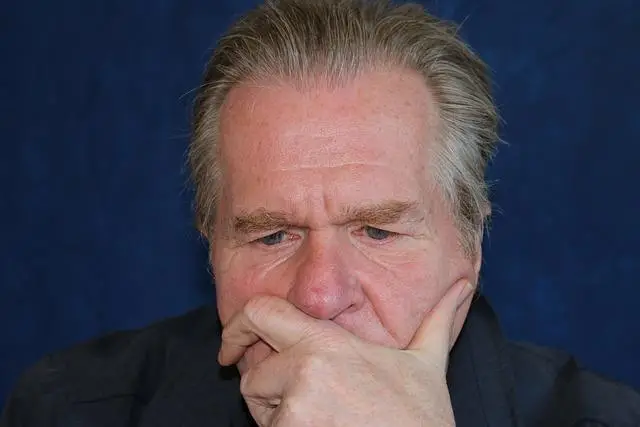 An older white man with square forehead and hair combed straight back, head resting on his hand, thinking.
