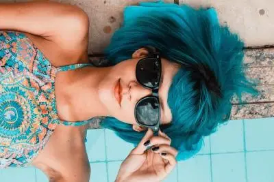 A young woman with teal blue hair and sunglasses lying on the deck of a pool.
