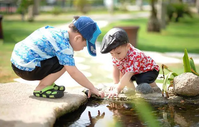 Two young boys squatting down, exploring a pond in a park.