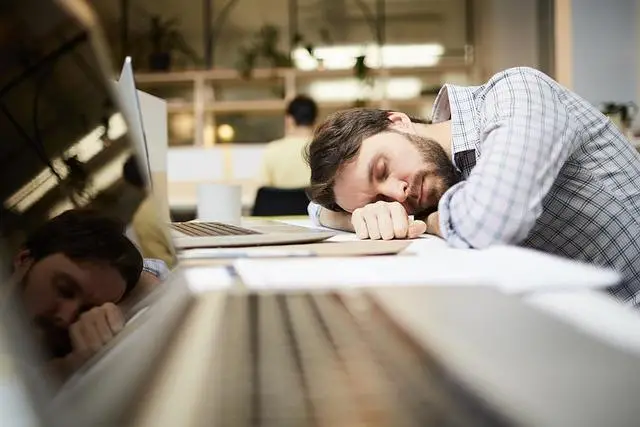 A young man with beard and moustache, head on his arm in front of his laptop, asleep.