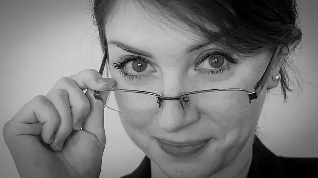 A black and white close-up of a middle-aged woman looking over the top of her glasses with a smirk.