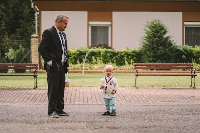 An older man and a toddler standing on a walkway, the older man looking down at the child.
