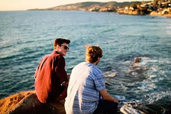 Two young men talking on a rock by the ocean.